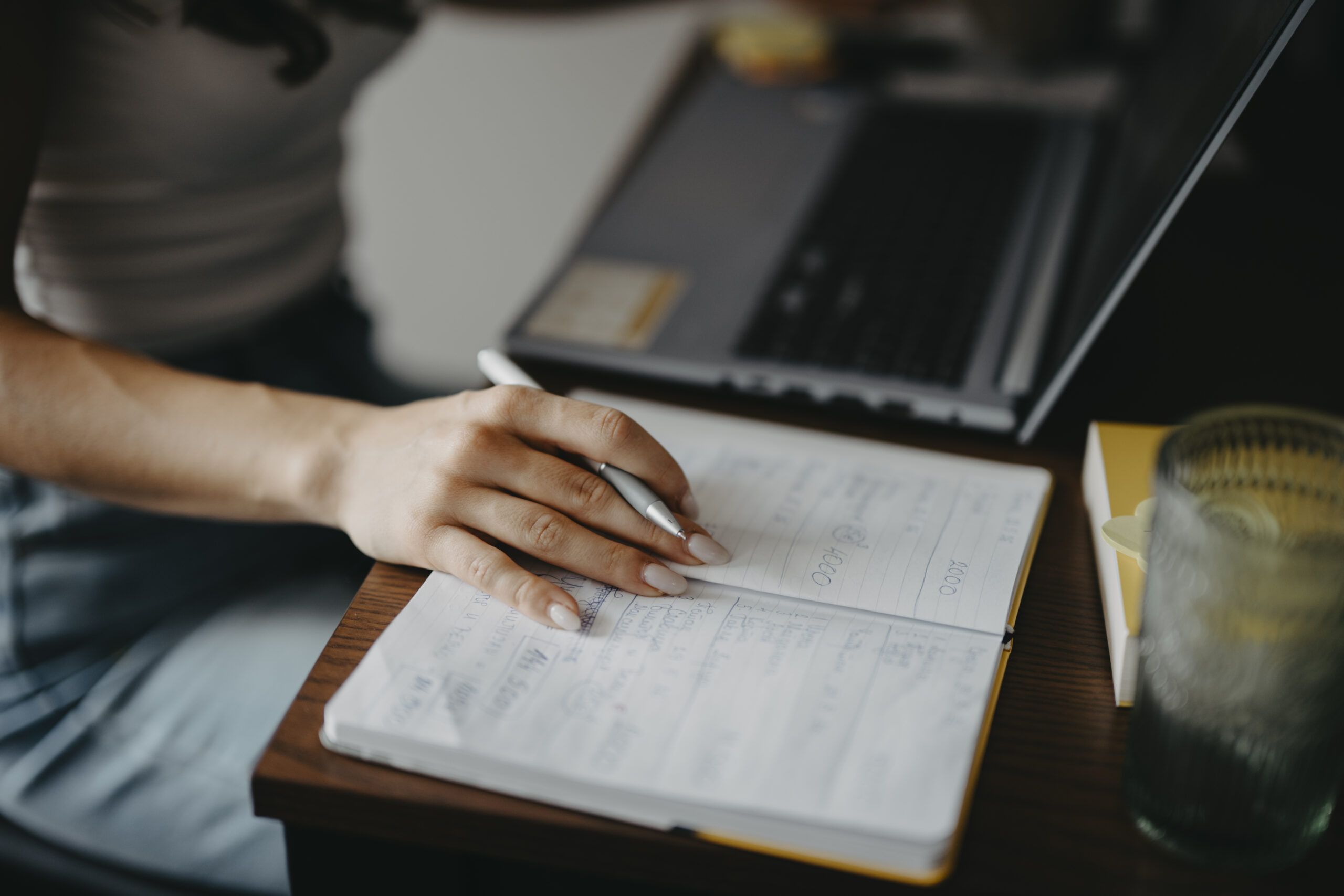 Person taking handwritten notes next to an open laptop at a desk